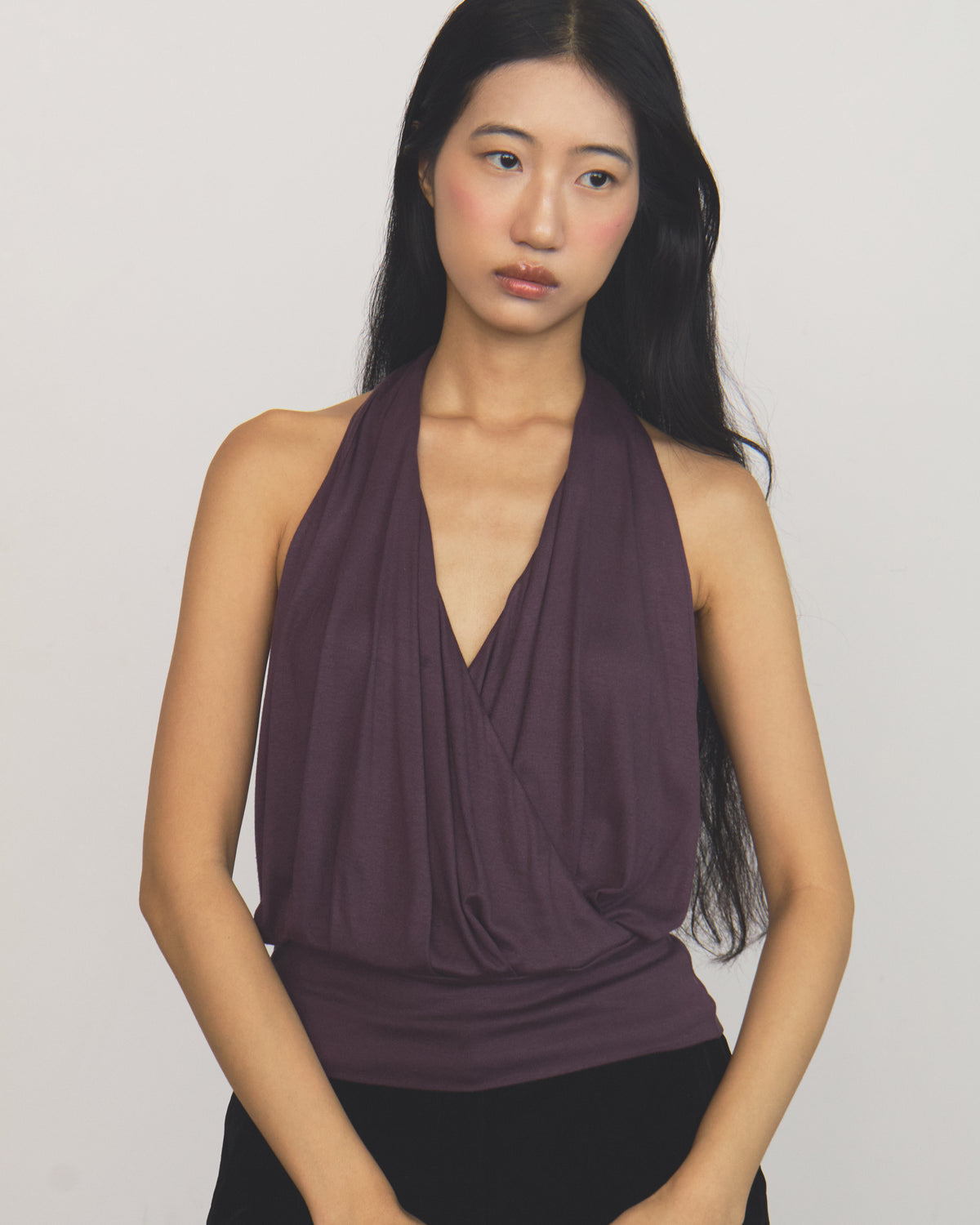 A woman modeling a sleeveless halter-style top with a draped wrap-front design in a deep plum purple color, paired with black bottoms, against a plain light gray background.